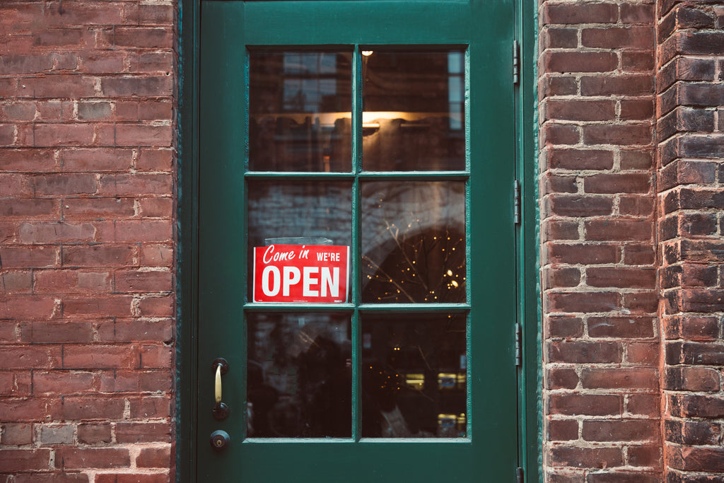 A red open sign hangs in the window of a green door to welcome people into the store.  Brick building holds up the storefront where a merchant sells clothing, hats, accessories, shirts, coffee mugs, phone cases, art, downloads, music, books, lesson & subs