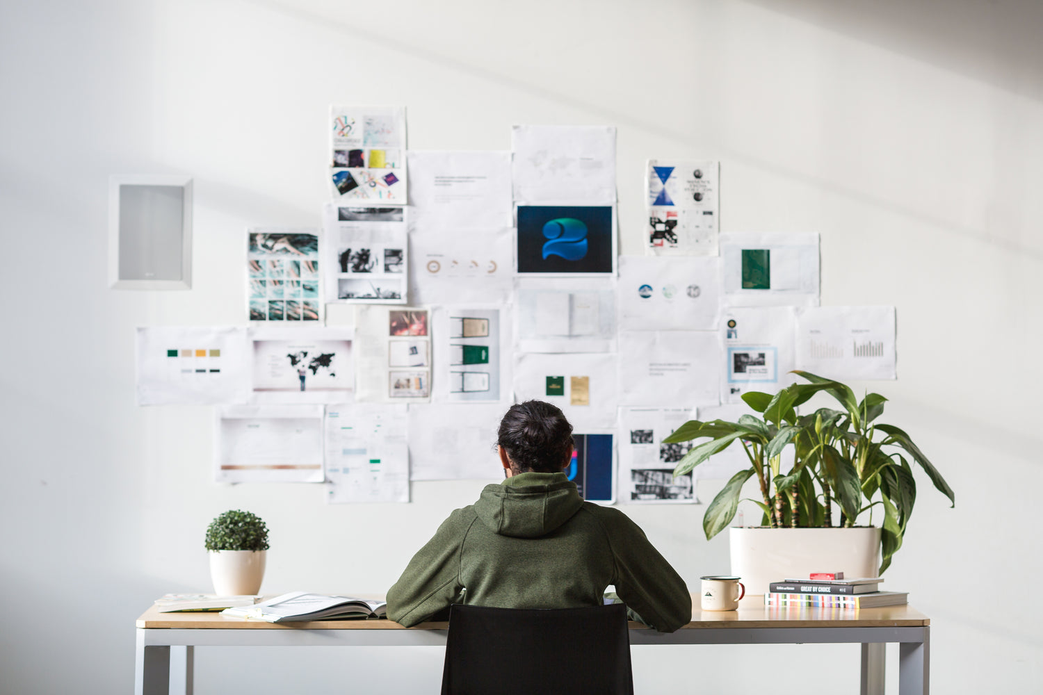 Marketing person sits at desk.  Sketches on wall of designs, ideas, pictures, moodboards, examples, strategy, video concepts, content calendar.  A plant sits on the desk. Person writes copy, words, scripts and storyboards for production and delivery.